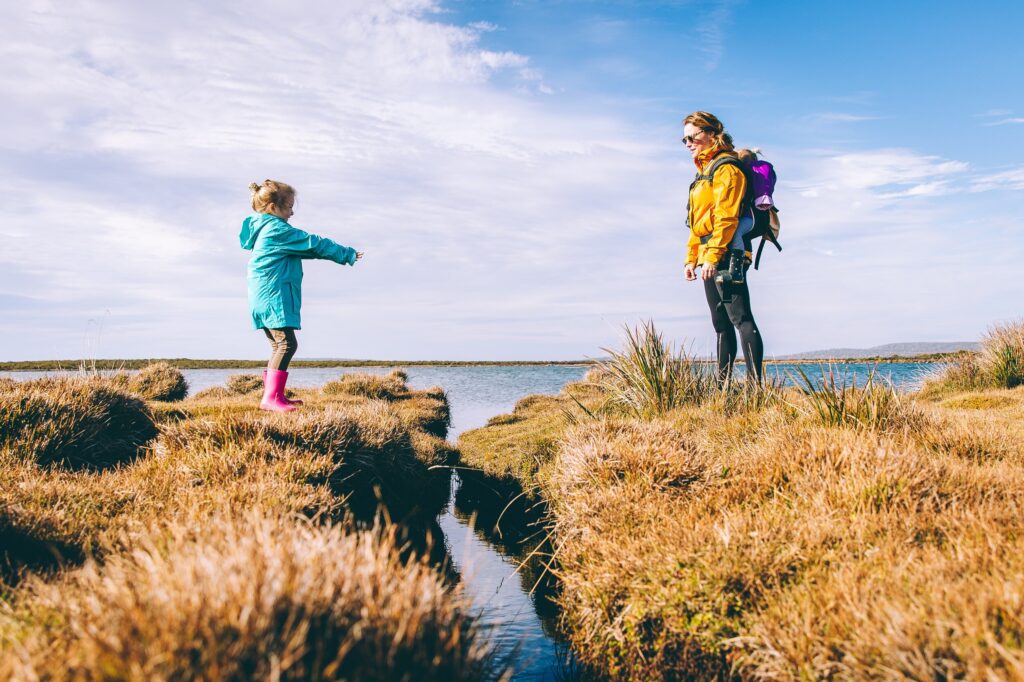 Photo of two children on a grassy shorline. The younger one is reaching across the water-filled ditch that separates them.