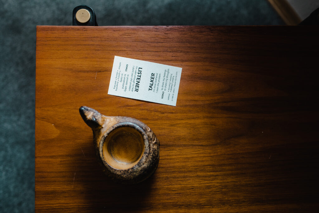 Photo of a small card and handmade coffee cup sitting on a wooden table. The card has the words "listener" on one end and "speaker" on the other.